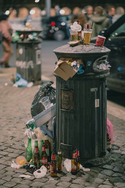A large pile of mixed household waste and rubbish overflowing from multiple clearly visible bins on a paved urban street. The contents include cardboard boxes, paper bags, plastic packaging, black and