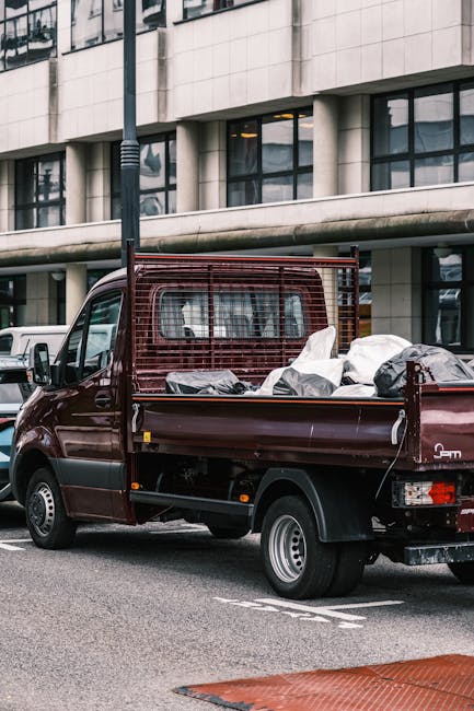 A dark red flatbed utility vehicle, parked on a city street in front of a modern building with large windows and concrete facade, is loaded with various discarded items including black and white bags,